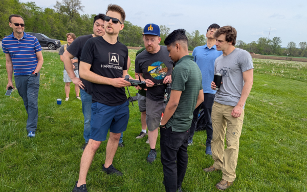 A group people standing outdoor in a field, one holding a control and talking.