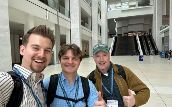 Three smiling people pose for a selfie in a spacious convention center lobby.