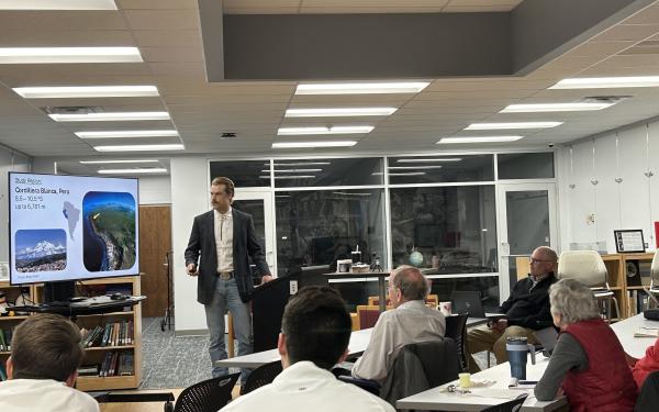 A presentation with an audience listening to a speaker standing next to a screen.