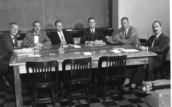 A black and white photo of a group of men in suits sitting around a table with many documents strewn about.