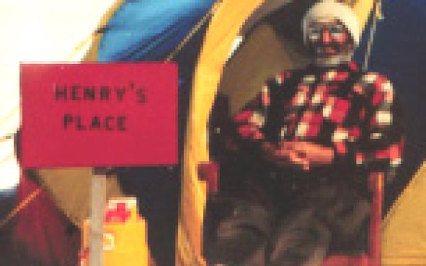 A white man wearing a flannel, hat, thick boots, pants, and a pair of sunglasses lounging in a wooden chair. He is sitting in front of a tent. A sign next to him reads "Henry's Place"