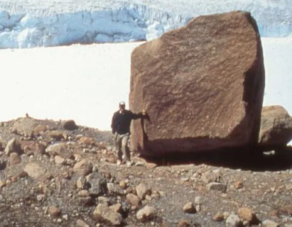 A person standing next to a large boulder in a rocky terrain with snow and ice in the background.