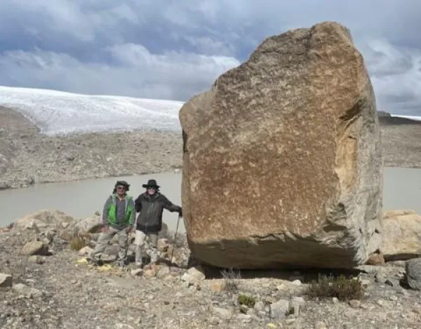 Two people standing next to a large boulder in a rocky terrain with snow and a lake in the background.