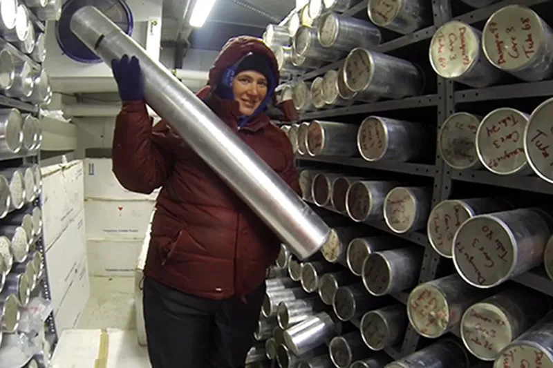 A scientist holding an ice core cyllander inside the ice core storage freezer. There are dozens of cyllanders stored on the shelves.
