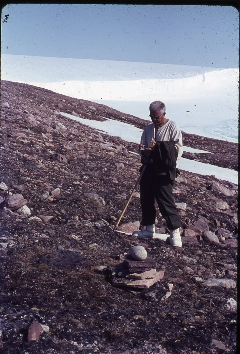 A man wearing glasses with a stick standing in a rocky mountainous terrain writing in a notebook he is holding and there is snow and a snowy plateau behind him.