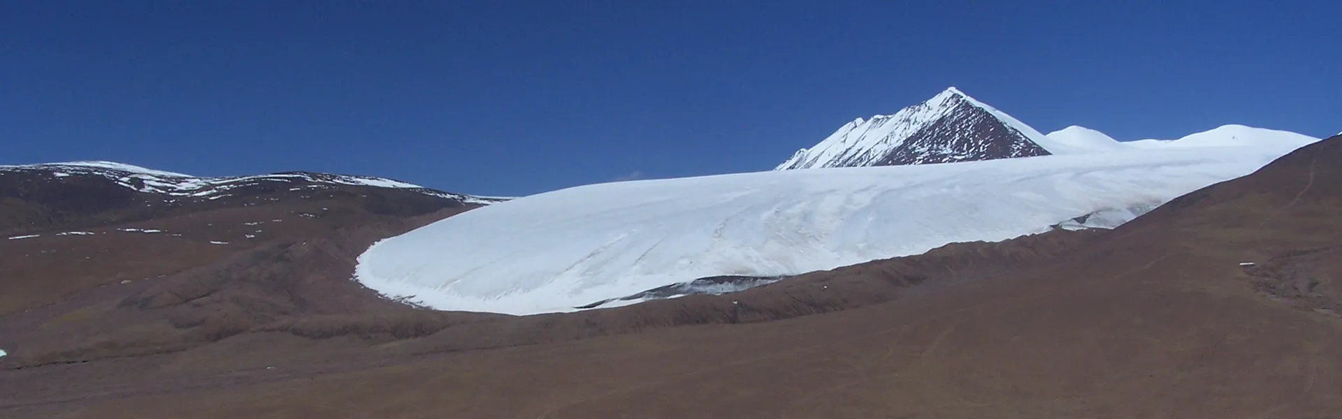 Landscape showing a mountain covered in snow in the distance. The foreground of the photograph reveals brown ground, not covered in snow.