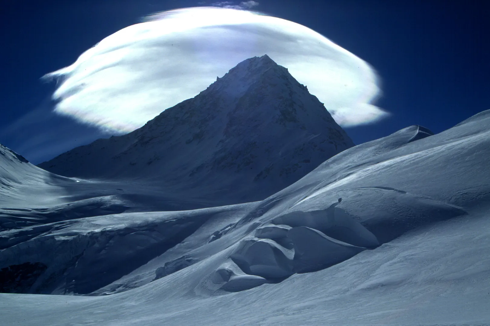 Snow covered ground with a mountain peak in a distance against a large white cloud under dark blue skies.