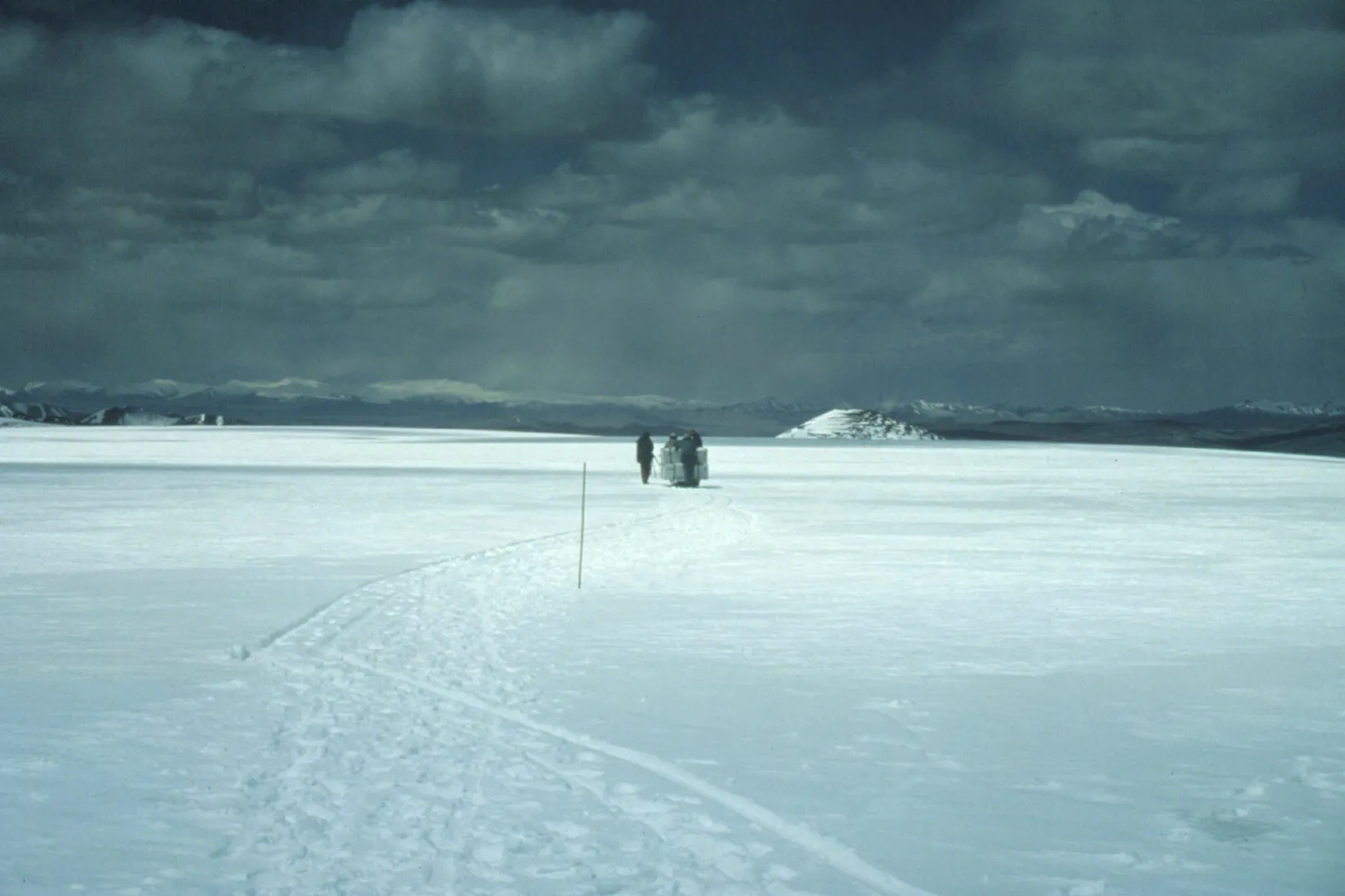 People walking in a distance in a snow covered field under gray, cloudy skies.