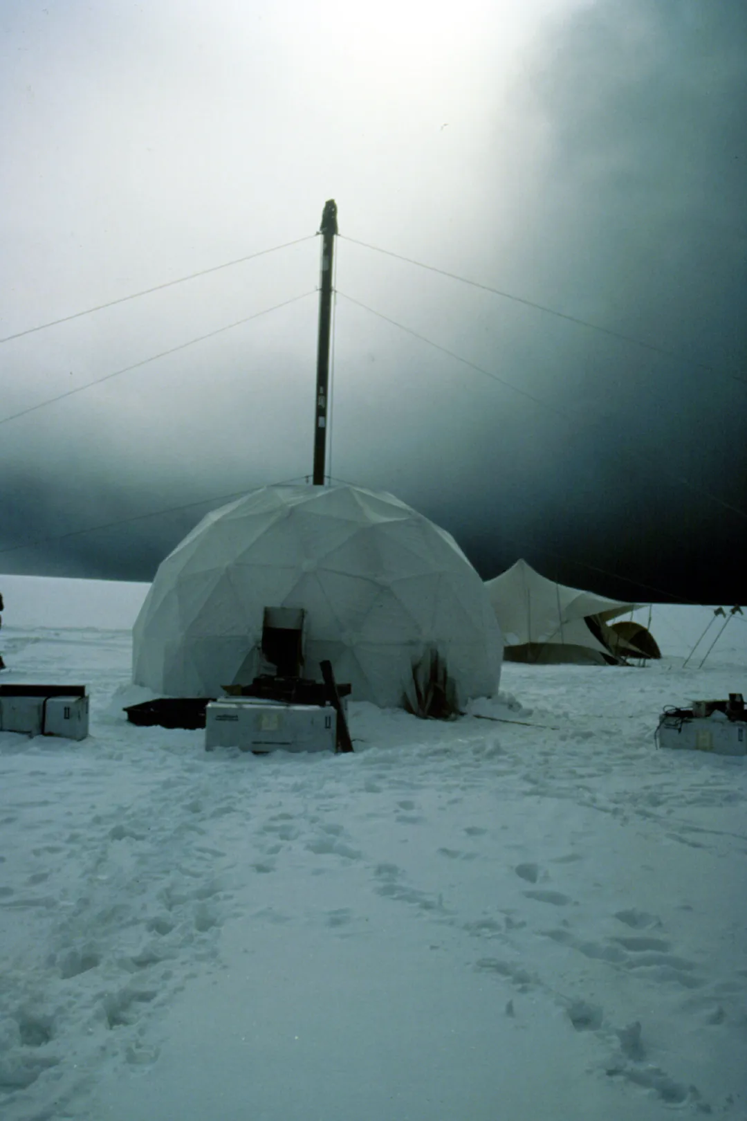 A dome looking tent with a pole sticking above it on snowy ground with white gray skies.