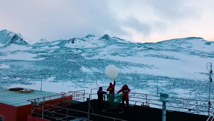 3 people standing on what seems to be a rooftop with a balloon with mountains in the backdrop