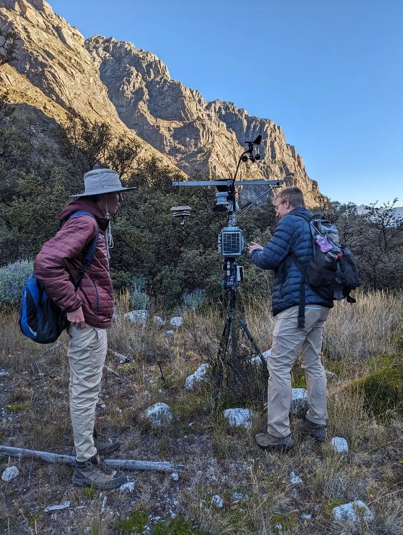 Two people standing in a rocky field in the mountains, working on equipment on a tripod.