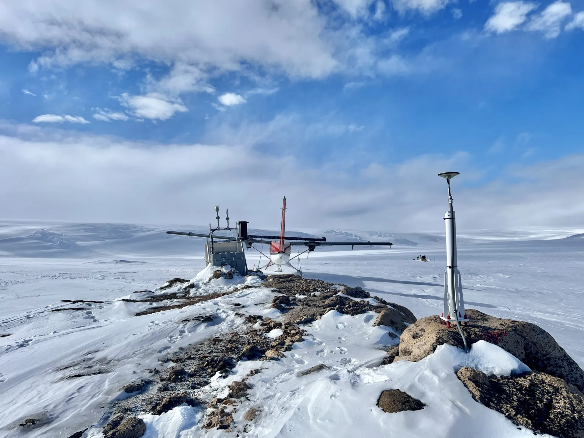 A vast snowy field with some rocks, equipment and a plane in the foreground under blue skies with white clouds.
