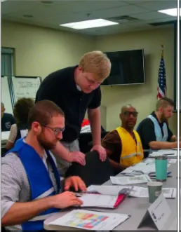 Alex McCarthy leaning on a chair looking at papers on a table in front of one of the seated people in a room. 