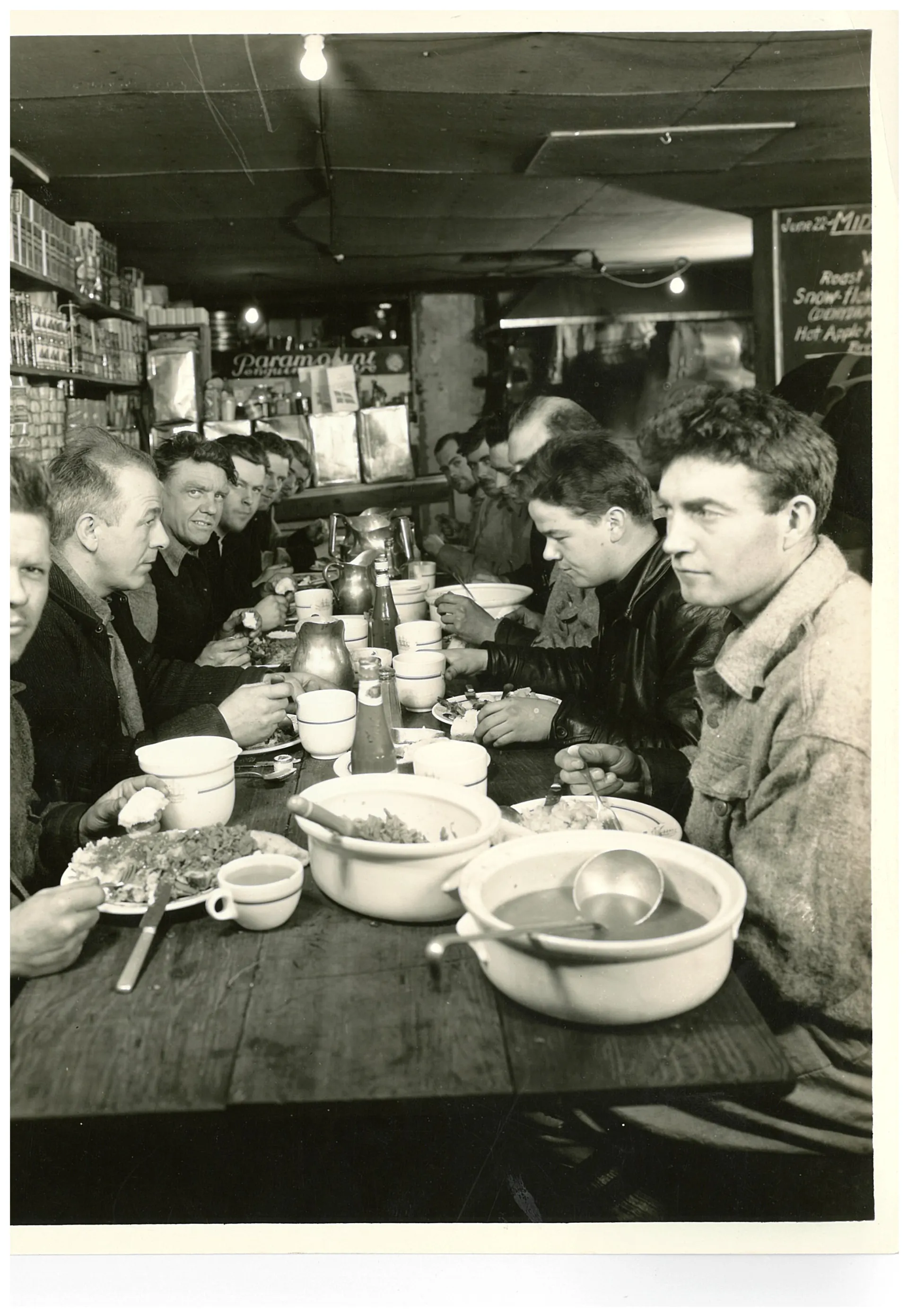 12 collective men seated before a buffet streching out across a wooden bench. A cutoff menu in the top right corner indicates the setting as a restaraunt.