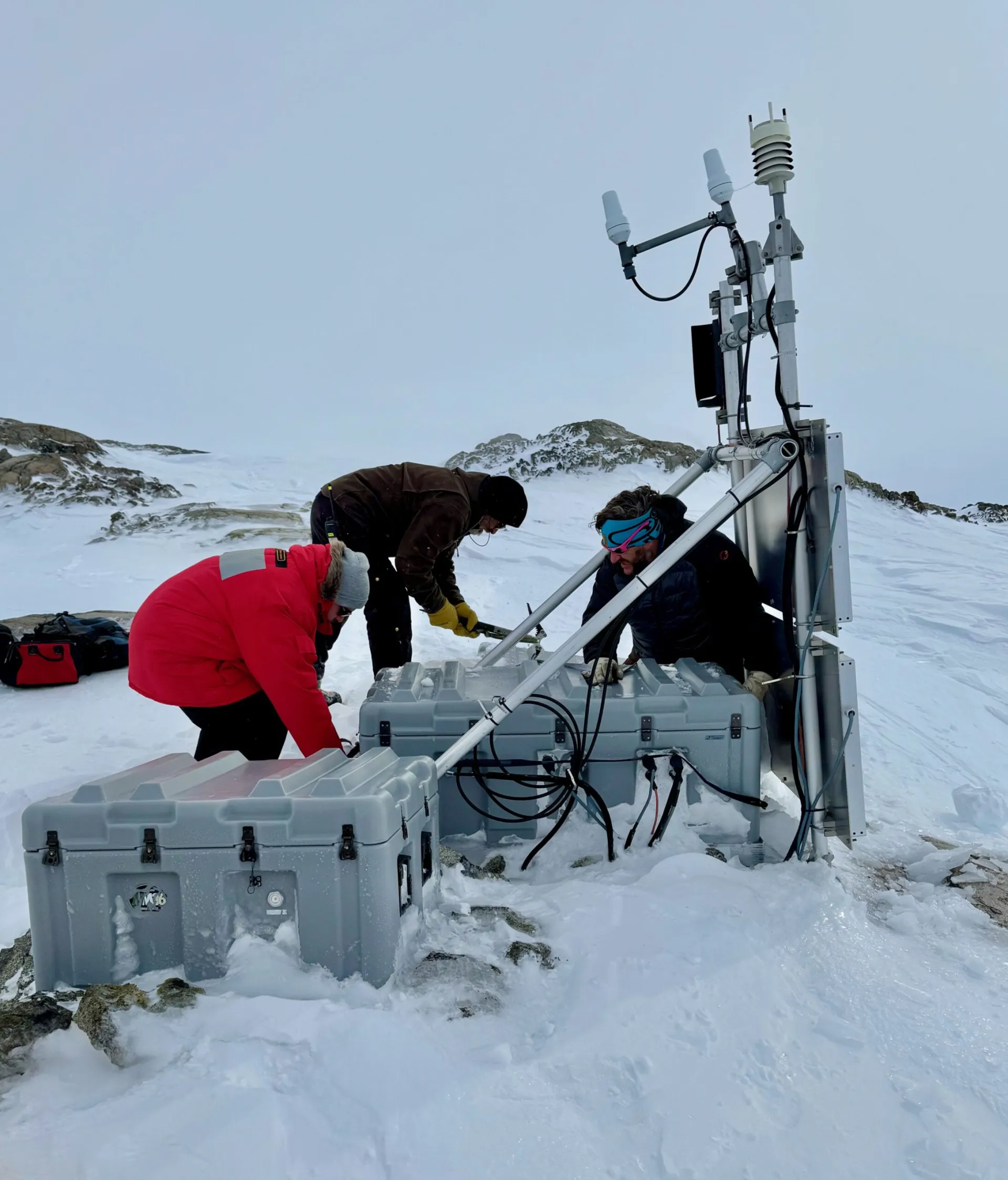 Three individuals inspecting scientific equipment on a snowy mountain.