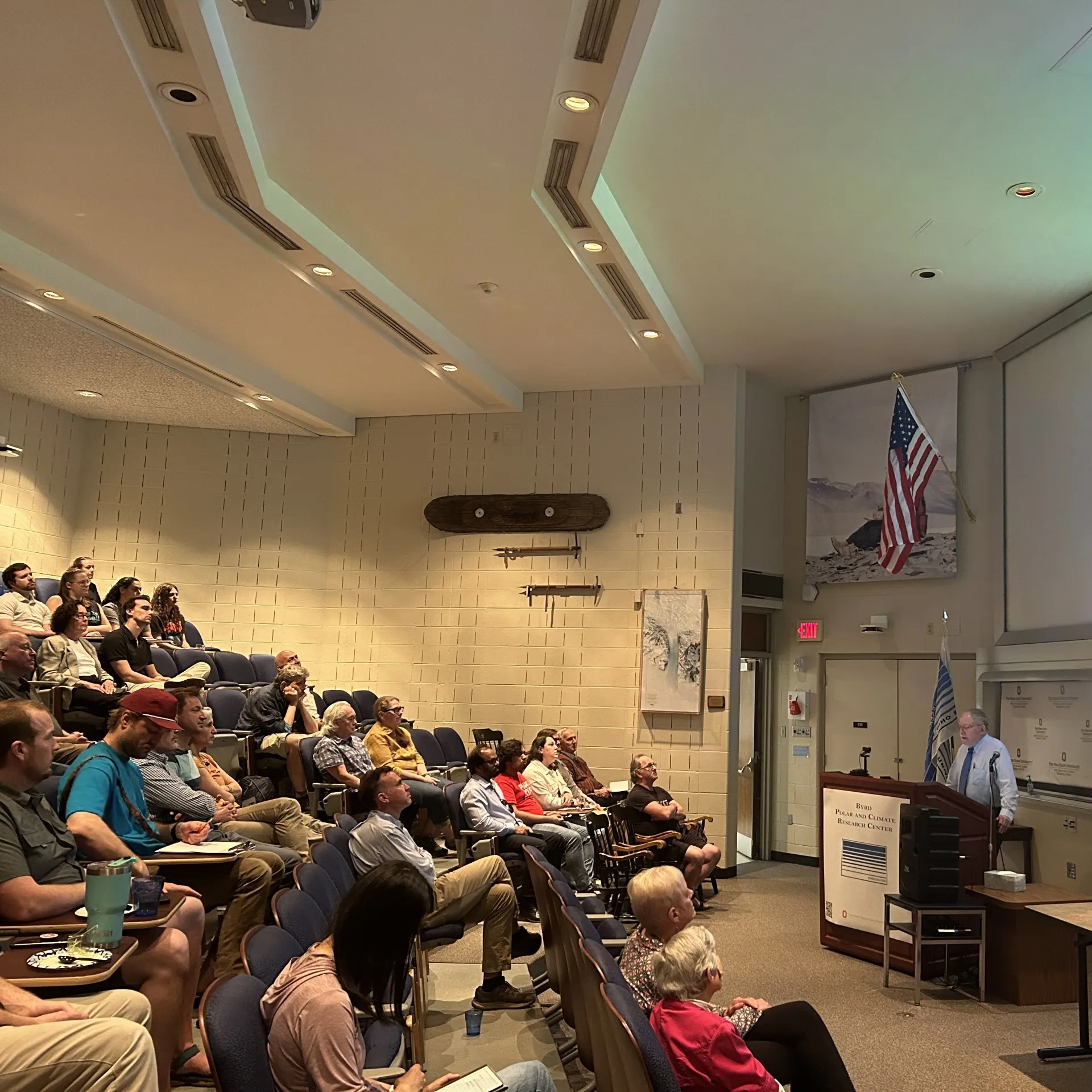 Audience seated in a lecture hall listening to a speaker at a podium, with a canoe mounted on the side wall.