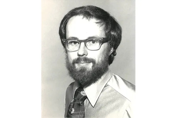 Black and white portrait of a man with glasses, a full beard, and short hair, wearing a collared shirt and a tie.