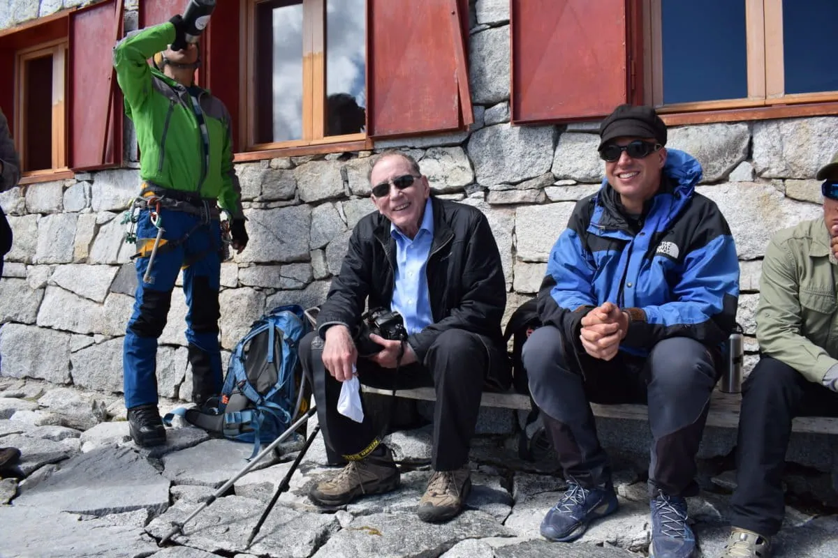 Two people sit smiling outside a stone mountain hut, with climbers and gear nearby.