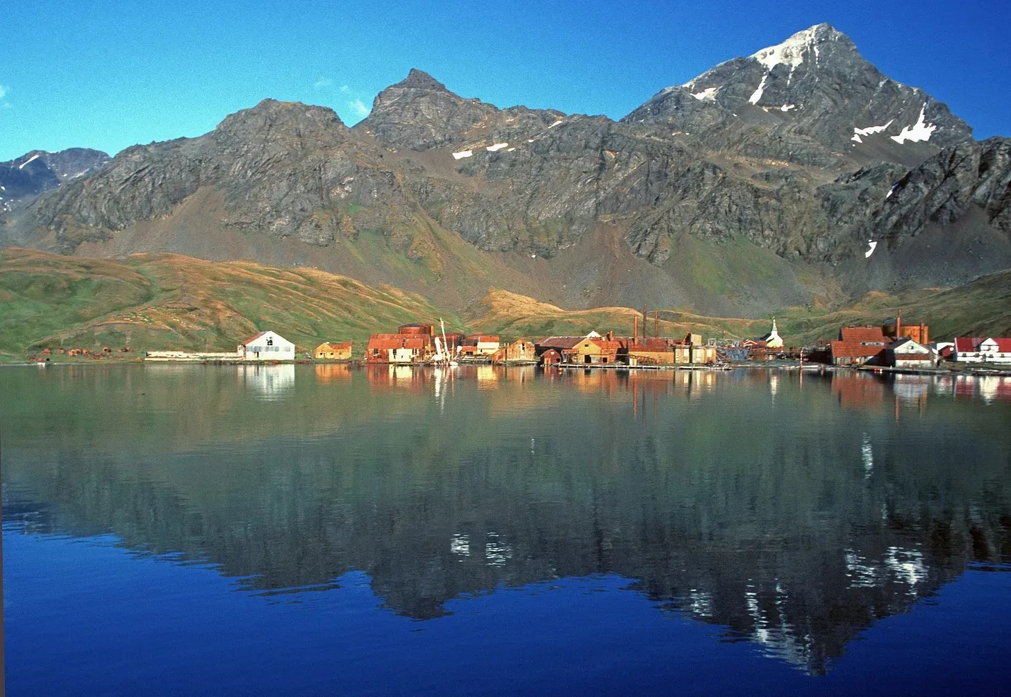 a coastal settlement with red-roofed and white buildings along the water’s edge, backed by steep rocky mountains with patches of snow near their peaks
