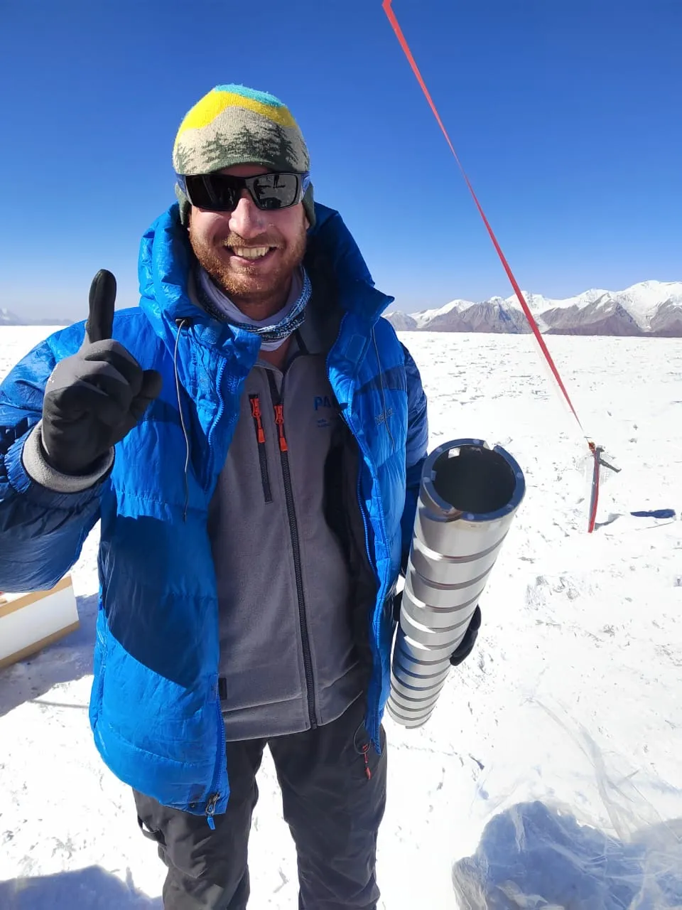 Smiling researcher on snowy glacier holding an empty ice-core barrel.