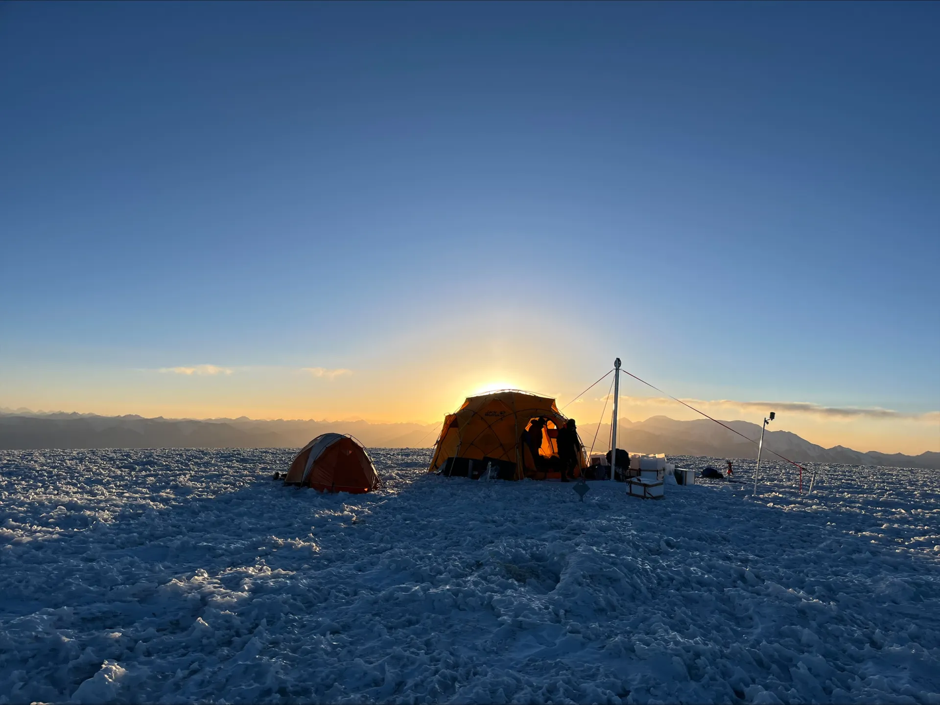 Glacier-top camp with lit tents under low-angle light near the horizon.