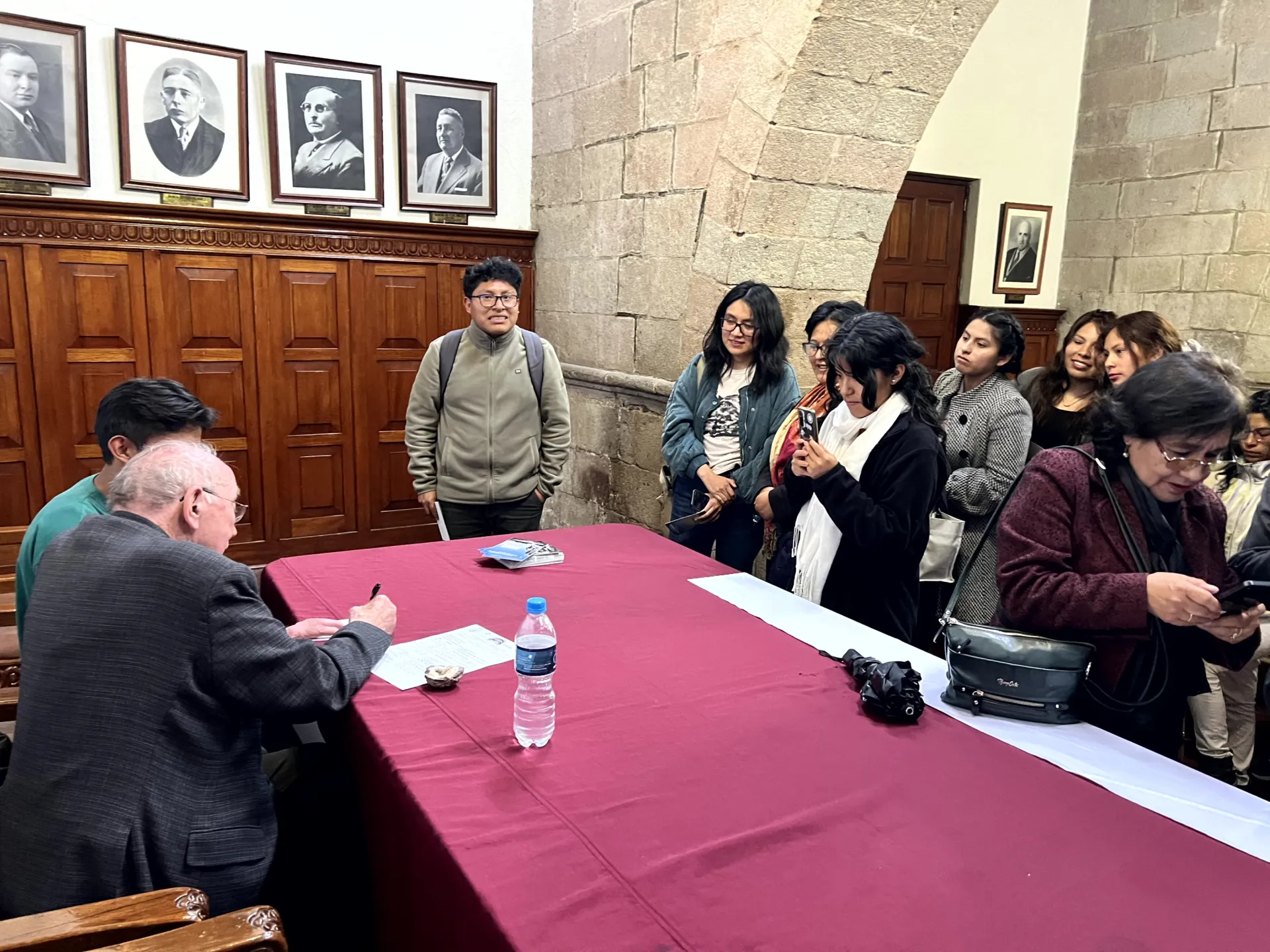 a person signs papers at a table as crowd gather in a historic university hall.