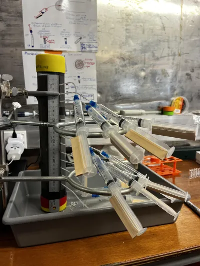 A number of syringes with clear liquid attached via clear tubes to a cylinder column with a ruler attached to it in a short plastic tub on table  in a lab