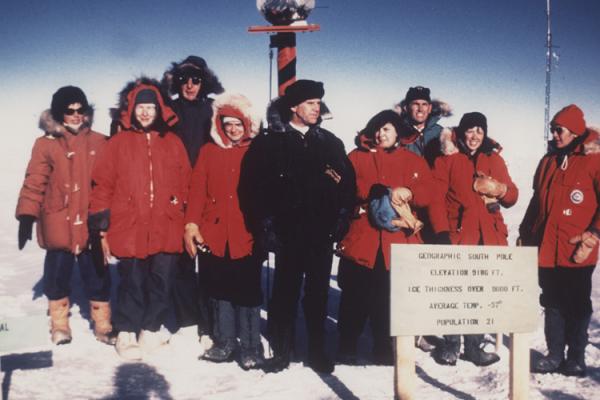 A group of women stand to take a photo at the south pole. The majority of the group is wearing heavy red coats. There is one individual in the center wearing a black coat.