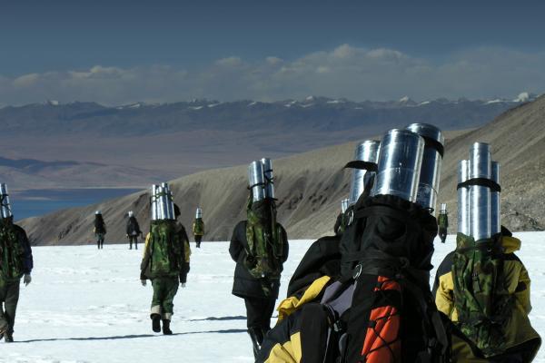 A group of people walking on a snowy ground and brown mountains in a distance, with silver cylinders on their backs.