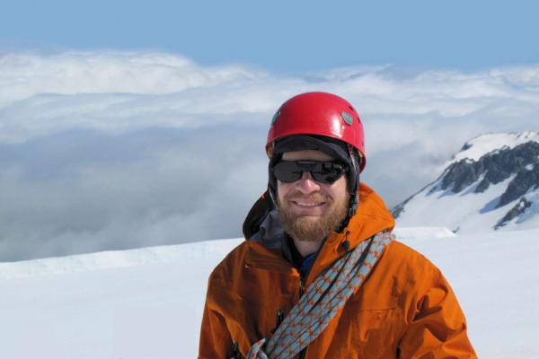 Richard Alley standing on a snow covered mountain. Richard is wearing a helmet and an orange coat with a length of rope hung across his chest