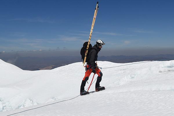 An indvidual wearing sold weather gear, a helmet, and research supplies hikes up a mountain in the arctic 