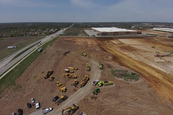 Sky view of freeway, construction site, equipment, vehicles, building and houses at a distance.