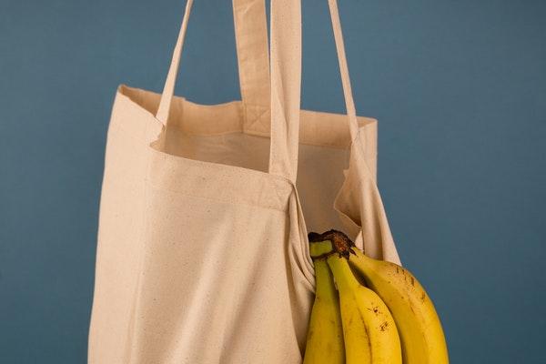 a hanging tan fabric shopping bag with a banana bunch hanging out with a gray-blue background