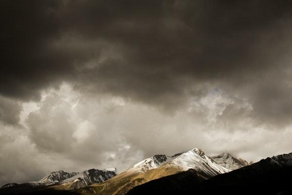 Snow covered, some treed and some brown mountains at a distance with dark cloud at the top but white clouds right about the mountains and dark mountains in the foreground.