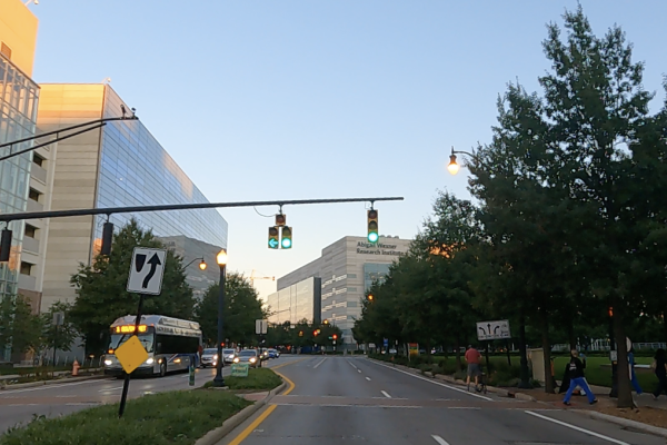A street view image of green traffic lights, with sidewalk, trees, buildings, bus and cars on the left, building straight ahead and green grass median and park, sidewalks and trees and people walking