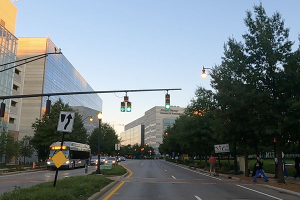 A street view image of green traffic lights, with sidewalk, trees, buildings, bus and cars  on the left, building straight ahead  and green grass median and park, sidewalks and trees and people walking