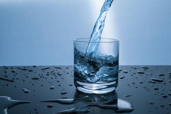 Clear water being poured from above into a short glass on a black shiny surface with water puddles and drops on it and a light blue background