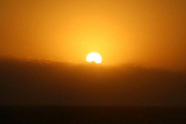 An orange sun partially behind puffy clouds with the whole image as orange, with the top half orange sky and the bottom half clouds and what seems to be land at the bottom as a darker shade of orangish brown.