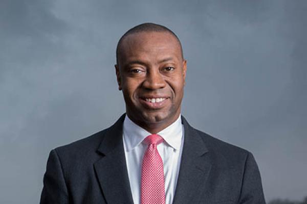 Portrait of Dr. J Marshall Shepherd with gray background smiling with very short hair wearing a white collar shirt and dark gray suit and pink rose tie and top and bottom of image have rose color