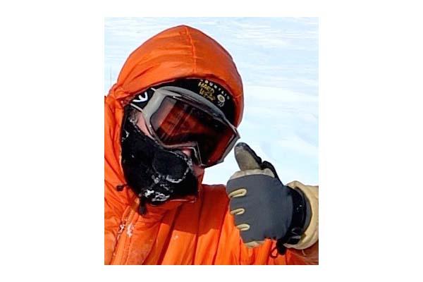 head and left hand and arm shot of Ian Howat wearing and orange buff jacket with a hood and wearing orange lens googles with gray frame and black mask covering mouth and nose giving a thumbs up wearing a black and tan glove with white clouds covering blue sky in background