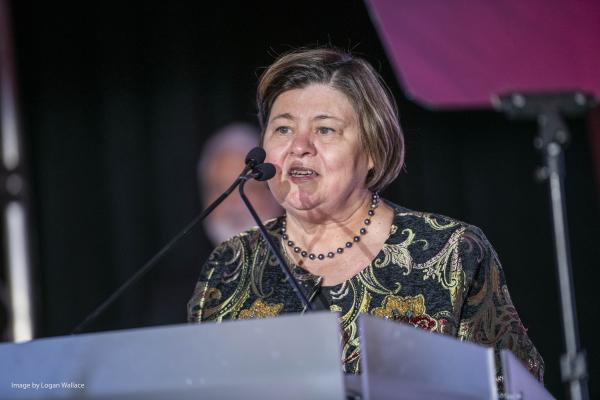 Georgie Shockey on stage indoor at a gray podium speaking with black background and purple lighting with short hair and gold black and red pattern jacket