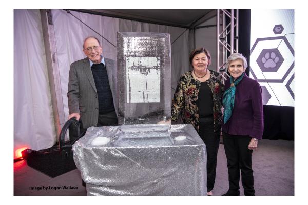 Left to right: Lonnie Thompson, Georgie Shockey and Ellen Mosley-Thompson standing in a room around a table with silver table cloth and a rectangular ice sculpture  on it with lilac colored carpet and white background with metal wires, and scaffolding