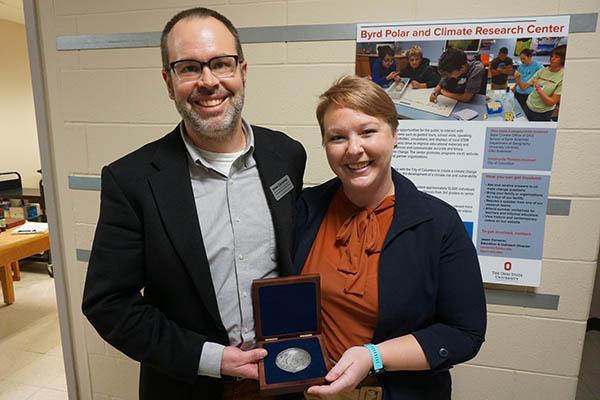 Jason Cervenec standing on the left side in the middle of the screen facing the camera with his right hand holding an open box with a round medal in it wearing a while collar shirt and a black blazer with glasses smiling while next to him Allison Chartrand is to his left holding that same box with her left hand wearing a black blazer and terracotta color silk shirt, light short hair and smiling