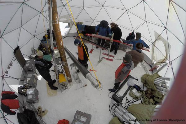 Image from above inside a core drilling dome with equipment around and scientists wearing winter clothes working on cylindrical long ice cores with snow on the ground
