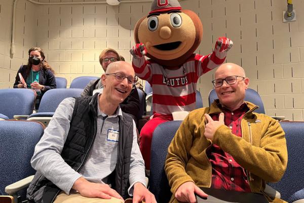 Matt Saltzman, with glasses wearing a light color long sleeve collar shirt and a dark gray vest on the left seated in auditorium blue cushion seat with Bryan Mark wearing glasses , a red and black flannel and a mustard color sweater on the right both smiling and Brutus in the middle and a lady wearing a mast in the far left upper sear and a man's head behind then