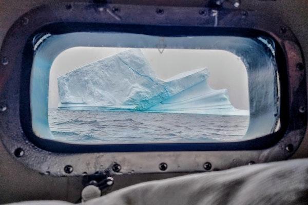 image of a a white sky and a blue white iceberg in the calm light blue water filling the porthole of a ship we look through with an elongated oval shape frame and rivets around the metal  frame