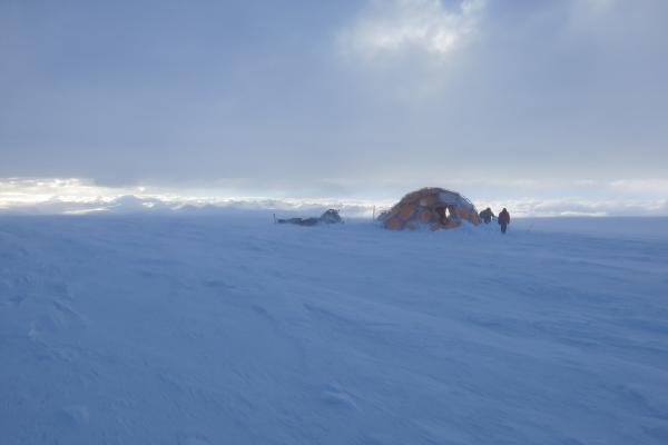 Snow Storm at Guliya ice cap drill site in 2015. Flat snow covered vista with grey clouds with some sunlight and blue skies at a distance illuminating a series of snow covered mountain peaks while in the foreground on the right side of the image there is an orange gray bubble tent with three people two working on something and one standing posing with a red coat