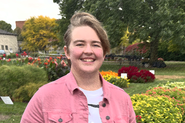 Standing in a garden is a white woman with short blonde hair wearing a pink jacket