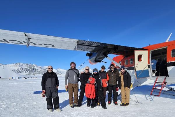 Several people posing next to a plane on a sunny day with blue skies in winter gear with snow covered mountains in the background and snow on the ground.