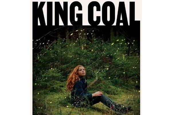 A person sitting on grass in the woods surrounded by shrubs with large text saying King Coal